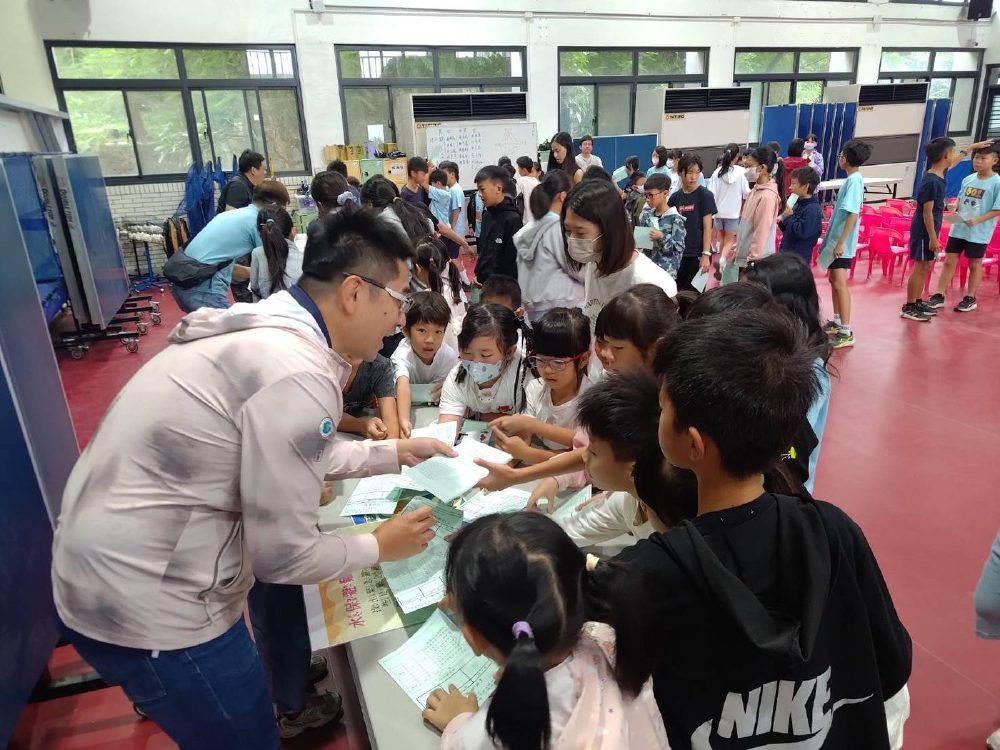 技師俯身在長桌前發下闖關學習單，一群小學生圍著桌子伸手接過，背景是排隊等候的同學。