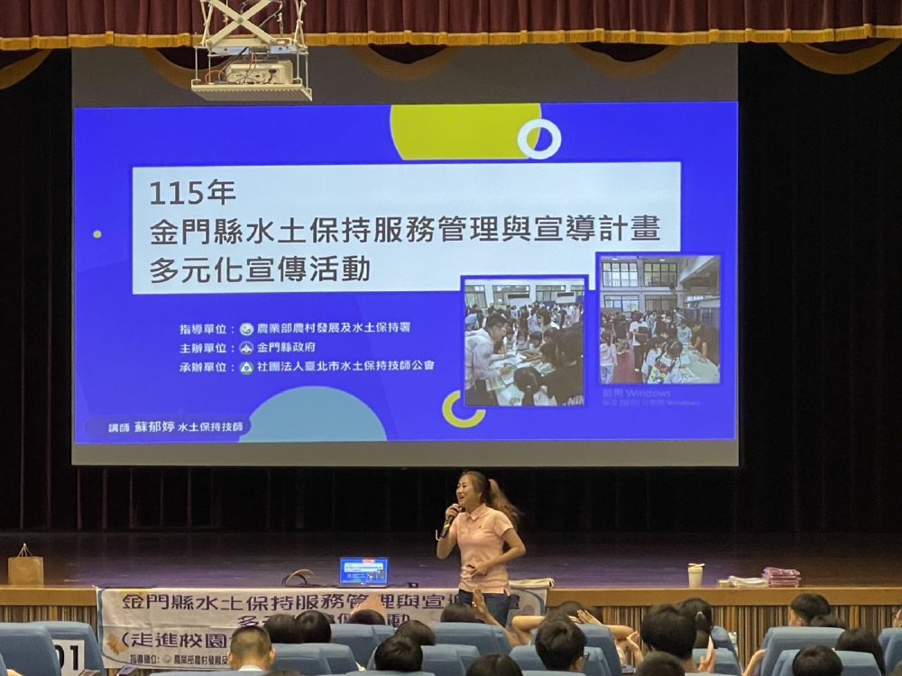 技師站在禮堂講台上拿著麥克風宣導，身後投影幕顯示「115年金門縣水土保持服務管理與宣導計畫多元化宣傳活動」簡報首頁，台下坐滿學生。